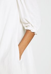 Bras d'une personne portant une robe blanche avec des manches à poignets élastiques, main placée dans la poche latérale de la robe.