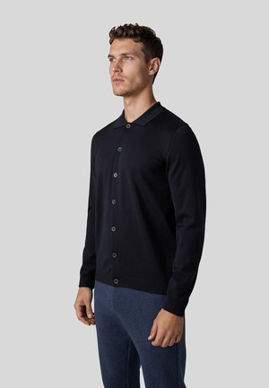 Mand iført en sort knapfast cardigan og blå bukser, stående og kigger let mod venstre mod en ensfarvet grå baggrund.