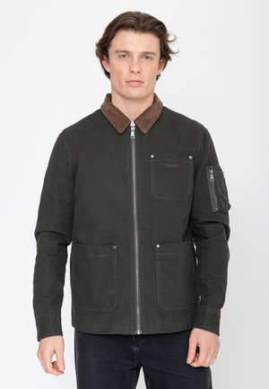 Jonge man draagt een donkergroene jas met ritssluiting, een bruine corduroy kraag en meerdere zakken aan de voorkant, staand tegen een effen achtergrond.