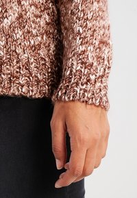 Brązowy sweter z dzianiny o fakturowanym wzorze, z prążkowanym mankietem. Materiał łączy ciemne i jasne odcienie, tworząc przytulny wygląd.