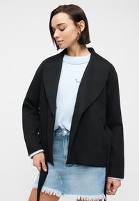 Femme portant un manteau noir ouvert sur un pull bleu clair et une jupe en jean effilochée, regardant vers le bas, avec des cheveux courts et foncés et des boucles d'oreilles anneaux dorés.