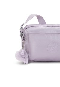 Lilakowa torba Kipling o gładkiej, błyszczącej fakturze, z zamkiem błyskawicznym i odpinanym breloczkiem w kształcie fioletowej małpki. Podkreślona naszywką z logo.
