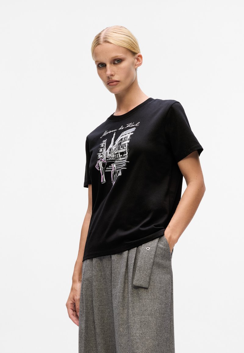 Blonde woman wearing black graphic t-shirt and gray wide-leg pants with hand in pocket, standing against white background.