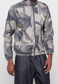 Mężczyzna ubrany w szarą i czarną, wzorzystą kurtkę na zamek marki Adidas oraz czarne spodnie, stojący na jednolitym tle.