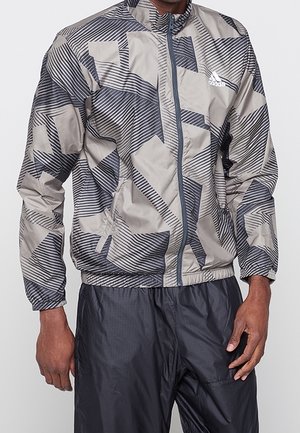 Uomo che indossa una giacca Adidas con zip grigia e nera a motivo, e pantaloni neri, in piedi davanti a uno sfondo semplice.