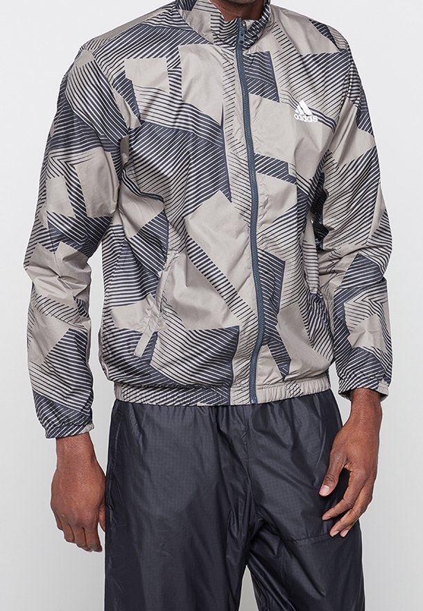 Mężczyzna ubrany w szarą i czarną, wzorzystą kurtkę na zamek marki Adidas oraz czarne spodnie, stojący na jednolitym tle.