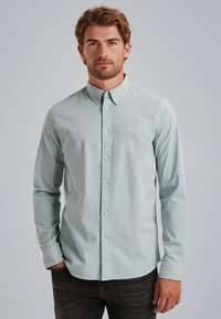 Camisa de botones azul claro con mangas largas, fabricada en suave algodón, y un pequeño logo en el lado izquierdo del pecho. Combinada con jeans gris oscuro.