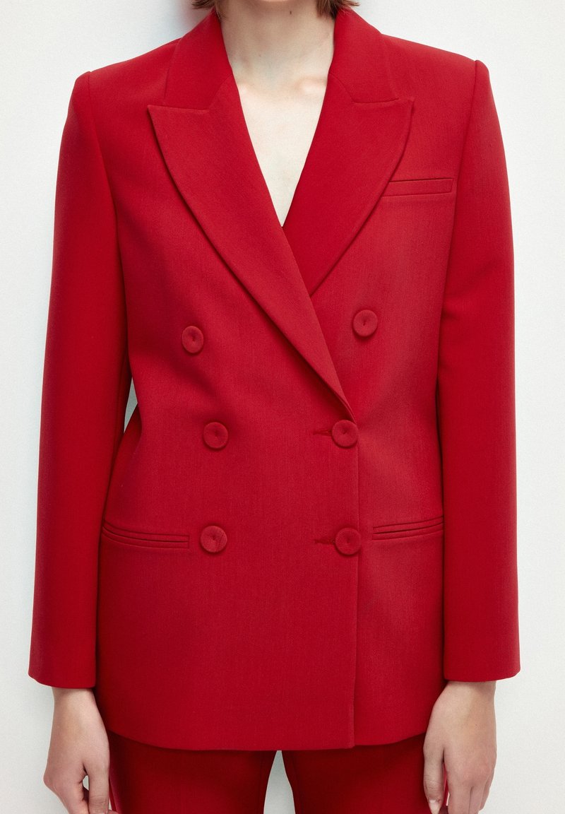 Femme portant un blazer rouge croisé sur mesure à six boutons et un pantalon rouge assorti sur un fond uni.