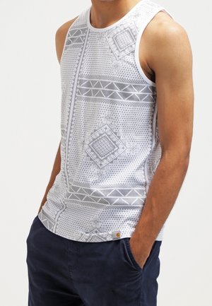 Mężczyzna ubrany w białą bezrękawnikową koszulkę z jasnoszarymi geometrycznymi wzorami i ciemnoniebieskie spodnie, ręce w kieszeniach.