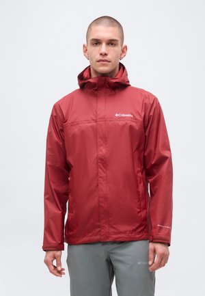 Hombre con chaqueta impermeable roja Columbia con capucha puesta, de pie frente a un fondo liso y claro, mirando hacia adelante.
