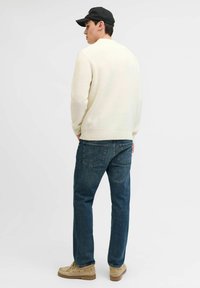 Suéter de punto beige, pantalones vaqueros azul oscuro, zapatos beige. Hombre de espaldas, con una gorra negra, se encuentra frente a un fondo blanco liso.
