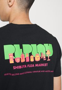 Camiseta de algodón negra con un atrevido texto gráfico en verde y rosa y ilustraciones de varios objetos en la parte trasera, incluyendo una lámpara y botellas.