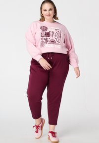 Sweat-shirt rose avec un motif graphique, associé à un pantalon de jogging bordeaux. Le modèle porte des baskets roses avec des lacets rouges. Textures lisses partout.