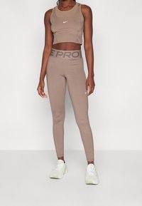 Top da allenamento a fascia corta di colore marrone abbinato a leggings a vita alta. Presenta un piccolo logo Nike bianco e un tessuto elastico e morbido.