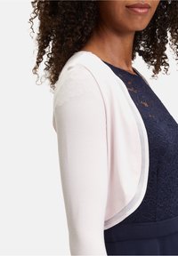 Bolero de punto color rosa claro con textura suave, con mangas abiertas y un diseño en capas sobre un vestido de encaje azul marino.