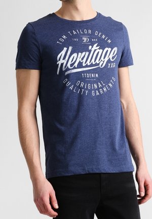 Mężczyzna w granatowej koszulce z krótkim rękawem z białym napisem "Heritage" i logo, w połączeniu z czarnymi spodniami.
