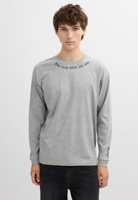 Camisa gris de manga larga hecha de algodón. Presenta texto negro en el escote: "Cosechas lo que siembras." Diseño simple con cuello redondo.
