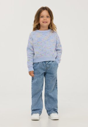 Maglione lavorato a maglia di un lilla chiaro con cannucciate multicolori, abbinato a jeans larghi in denim e sneakers bianche. Lo sfondo è bianco puro.