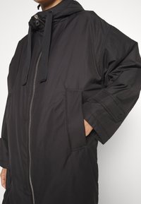 Manteau noir à capuche avec fermeture éclair, deux poches latérales et poignets élastiques. Confectionné en un matériau lisse et léger avec des caractéristiques de design minimales.