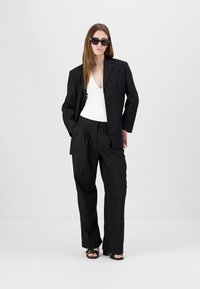 Svart dobbelspent blazer over en hvit topp, kombinert med løse svarte bukser og svarte solbriller. Glatt stoff, strukturert design.