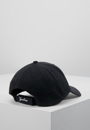 Czarna czapka bejsbolowa z regulowanym paskiem, haftowany napis "Yankees" w białym kolorze, umieszczona na białej narożnej powierzchni na tle szarego tła.