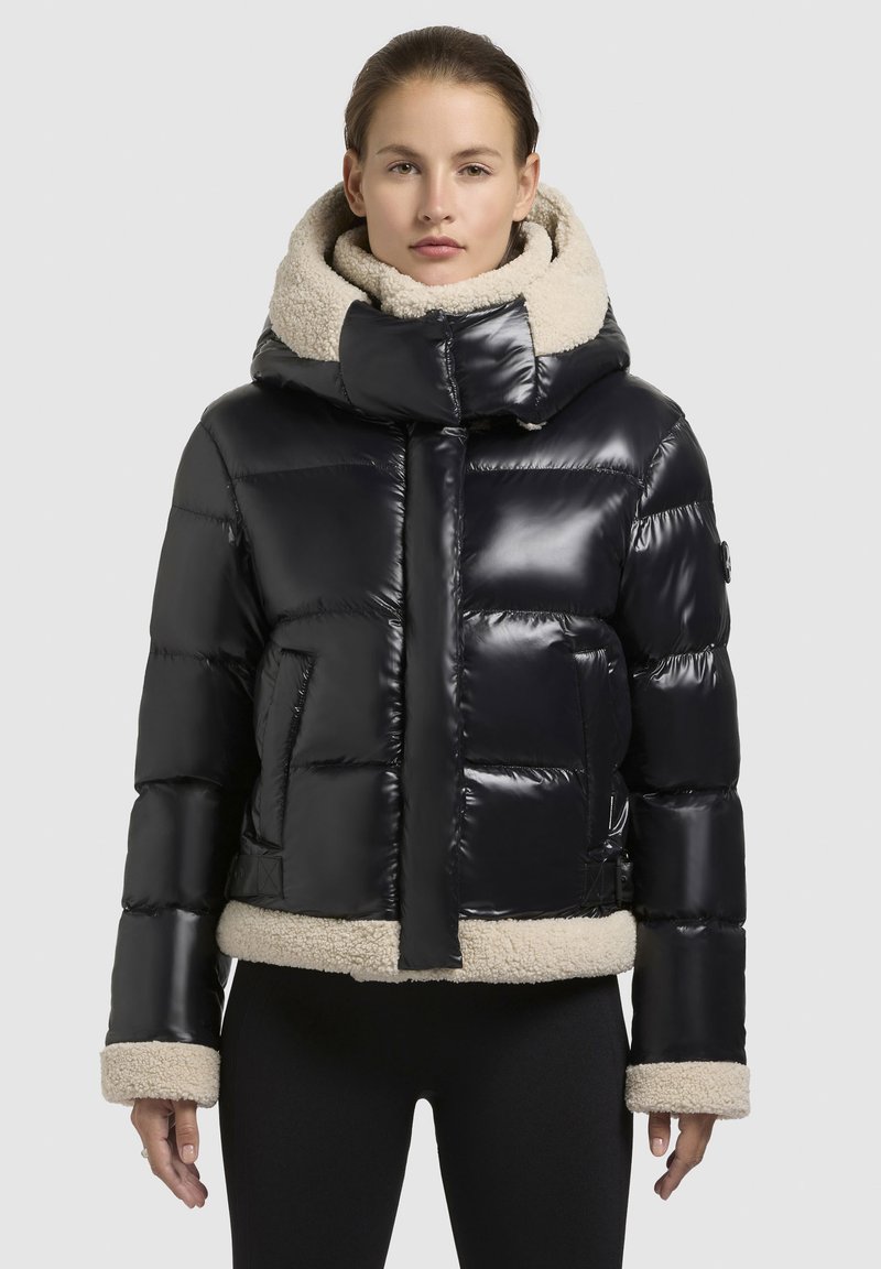 Zwarte gewatteerde jas met een glanzende afwerking, voorzien van een beige fleece-voering in de capuchon en onderrand. Het ontwerp heeft een ritssluiting aan de voorkant en zijzakken.