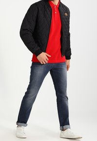 Schwarze gesteppte Bomberjacke mit gerippten Bündchen, rotes Polohemd, blaue Jeans und weiße Sneakers. Lässiges, geschichtetes Outfit mit auffälligem Farbkontrast.