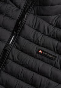 Chaqueta negra acolchada con cremallera central visible y logo de Superdry en parche y tirador de cremallera.