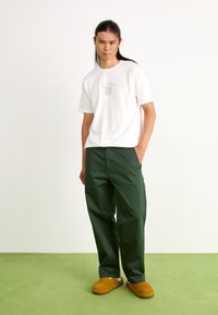 Tricou alb din bumbac cu imprimeu grafic, asortat cu pantaloni verzi închiși și saboți din suede maro. Fundalul este simplu, alb și verde.