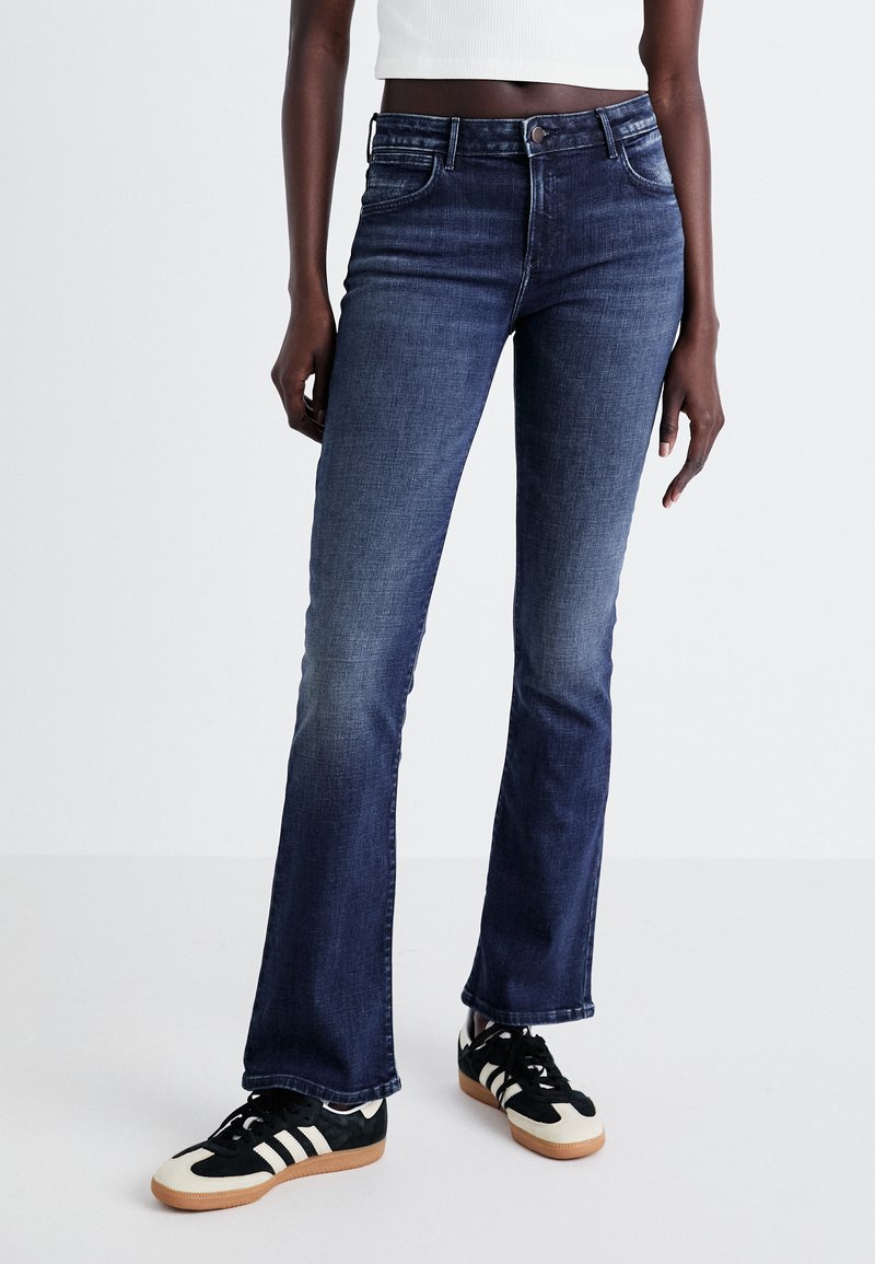 Jean évasé en denim de teinture foncée, présentant un design classique à cinq poches et un léger délavage. Associé à des baskets noires avec des rayures blanches.