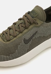 Sneaker verde oliva con tessuto a rete e dettagli testurizzati. Presenta lacci beige e un logo nero. Suola in gomma bianca con superficie a motivi.