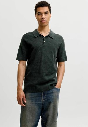 Homme aux cheveux courts et bouclés porte un polo en maille vert foncé à manches courtes et un jean bleu, debout avec une main dans la poche.