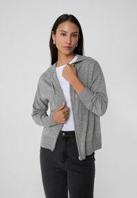 Sudadera gris con cremallera, hecha de una tela suave, que cuenta con una cremallera frontal, capucha con cordón y puños acanalados, llevada sobre una camiseta blanca.