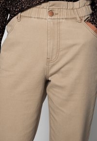 Pantalones de mezcla de algodón beige con una cintura ajustada, detalle de botón de cobre y bolsillos laterales. Textura suave y diseño de pierna recta.