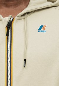 Κοντινό πλάνο μπεζ φούτερ με φερμουάρ, πολύχρωμο ριγωτό φερμουάρ, κορδόνι και μπλε-πορτοκαλί λογότυπο Κ-Way στο στήθος.