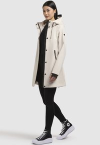 Manteau imperméable beige clair avec capuche, accents noirs, boutons-pression et poches latérales, associé à un legging noir et des baskets montantes.