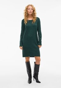 Abito verde a maglia con maniche lunghe, scollatura a girocollo e orlo a coste. Abbinato a stivali neri con tacco alti fino al ginocchio. Texture liscia e forma aderente.