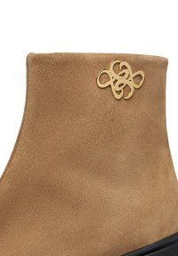 Stivale ankle in suede beige con suola nera, caratterizzato da un logo decorativo in oro a forma di anello intrecciato vicino al bordo superiore.