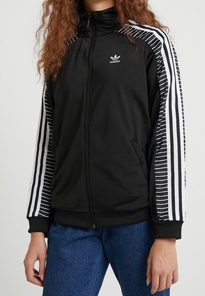Personne portant une veste zippée noire Adidas avec des rayures blanches et des manches à motifs, associée à un jean bleu, debout devant un fond uni.