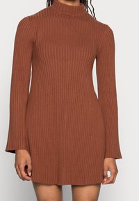 Robe marron côtelée avec un col haut et des manches longues, présentant une silhouette ajustée et des extrémités de manches évasées. Texture de tissu douce.