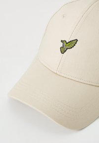 Beige baseballkeps i strukturerad tyg, med en grön och grå broderad kolibri på den vänstra frampanelen.