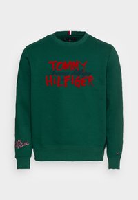 Felpa verde con collo rotondo, presenta un vistoso testo rosso e nero "TOMMY HILFIGER" sul davanti e il logo ricamato sulla manica sinistra.
