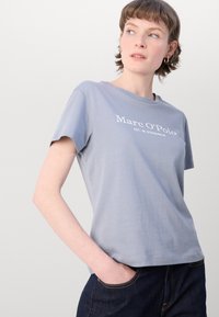 Svetle modré bavlněné tričko s krátkými rukávy, kulatým výstřihem a bílým potiskem loga "Marc O'Polo EST. V STOCKHOLMU." na přední straně.