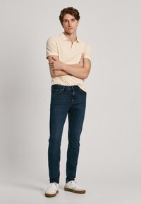Polo de color beige claro combinado con jeans azul oscuro. El modelo lleva zapatillas blancas con suelas marrones. Ajuste casual, con un diseño simple y sin patrones visibles.