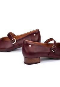 Zapatos de cuero burdeos con un tacón de madera bajo, que presentan una correa con una hebilla de plata y dos acentos metálicos en la parte trasera.