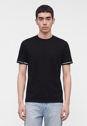 Jeune homme debout, face à l'avant, portant un t-shirt noir à manches courtes avec un texte blanc sur les bords des manches et un jean bleu clair.