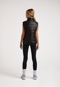 Schwarze ärmellose Steppweste mit hohem Kragen, mit vertikalen Steppnähten und einer glatten, glänzenden Textur. Getragen über schwarzen Leggings.