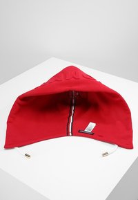 Capuche de sweat à capuche rouge avec cordons blancs et une bande verticale rouge, blanche et noire sur la couture centrale intérieure, posée sur une surface blanche.