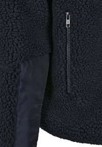 Veste en polaire noire avec tissu texturé, insert de manche noir lisse et une fermeture éclair argentée visible sur le devant.