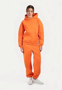 Oranje sweatshirt en broek outfit. Gemaakt van zachte stof, met capuchon en voorzak. Witte sneakers maken de look compleet.